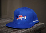 A royal blue flat-brim cap resting on a rustic wooden surface, featuring an embroidered fish design filled with an American flag pattern on the front and white text on the side.