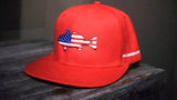 A bright red flat-brim baseball cap resting on a dark wooden surface. The front crown features an embroidered fish patch filled with an American flag pattern, and the side displays the white text 'PHILIPPEGAUT'.