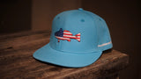 A light blue flat-brim cap resting on a rustic wooden surface, featuring an embroidered fish with an American flag pattern on the front and white text on the side.